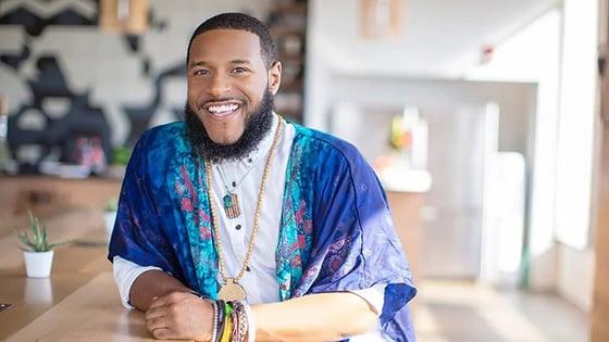 Rico Robinson stands in a café. He is wearing a blue-patterned wrap over a white button-up shirt. He is smiling.